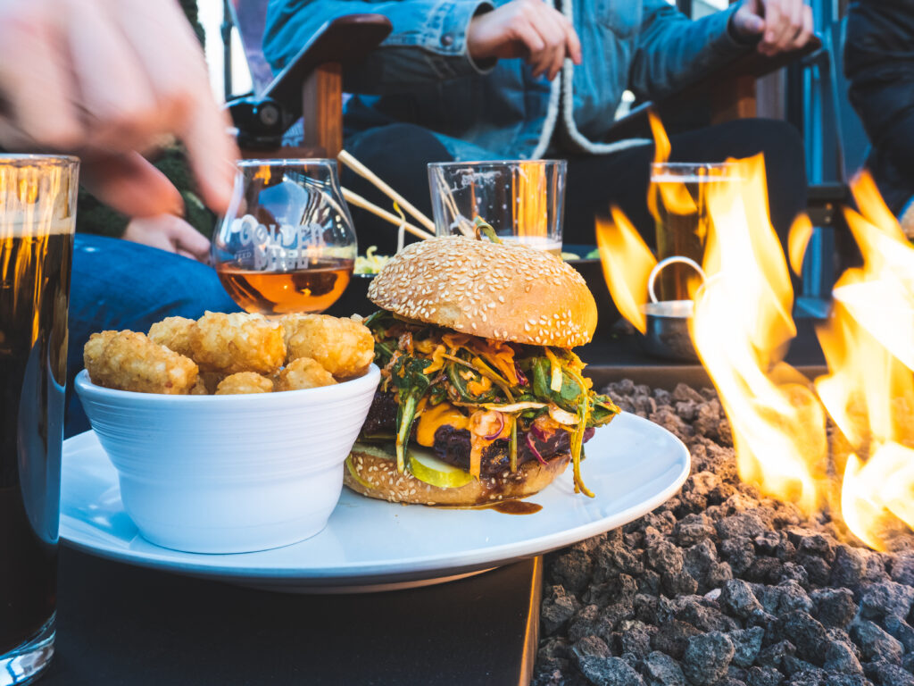 Friends gather around to enjoy a juicy burger and tater tots with beer next to a cozy fire at The Golden ill in Golden